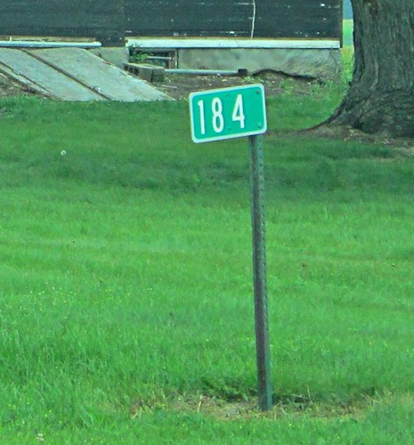 A reflective metal sign with an address of 184 mounted on a metal stake in a grassy yard with a house in the background
